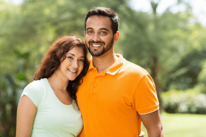 A man and woman smile joyfully together in a sunny park, surrounded by greenery and vibrant flowers.