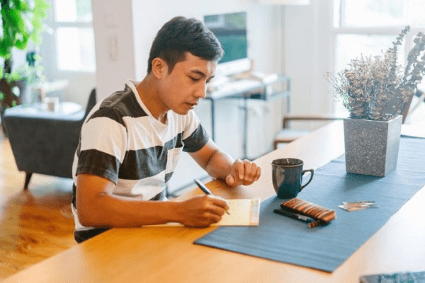 A man in a striped shirt sits at a table, writing in a notebook. A coffee mug, a bar of chocolate, and a vase with dried flowers are also on the table.